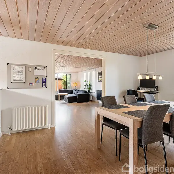 Spisebord med seks stole står i en lys spisestue med trægulv og træloft. I baggrunden ses en stue med sofaer og grønne planter. Et vindue viser udsigt til træer.