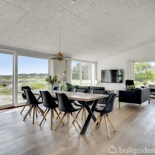 Spisebord med otte sorte stole står midt i et lyst rum med store vinduer. Stuen har en sofa, et fjernsyn og grønne planter. Udsigt til natur udenfor.