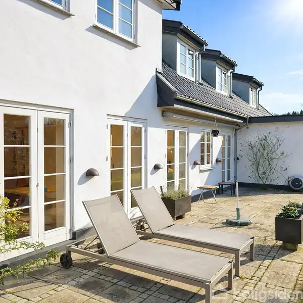 To liggestole står på en flisebelagt terrasse foran en hvid murstensbygning med flere døre og vinduer. I baggrunden er der grønne træer og en græsplæne.
