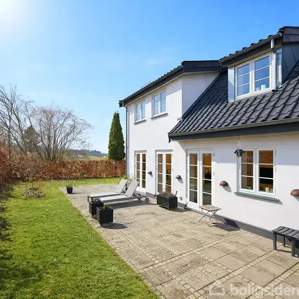 Hus med sort tag og hvide vægge står i en have med fliser, græs og liggestole. Solrigt vejr.