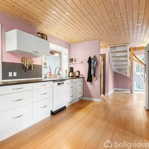 Køkken med hvide skabe og skuffer langs væggen, en opvaskemaskine, og sort bordplade. Trægulv og træloft i et lyserødt malet rum, med en trappe i baggrunden.
