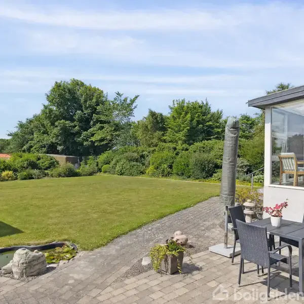 En terrasse med et sæt havemøbler står ved siden af et lyst hus med store vinduer. En velplejet græsplæne omgivet af buske fylder baggrunden, og der er et lille vandbassin nær terrassen.