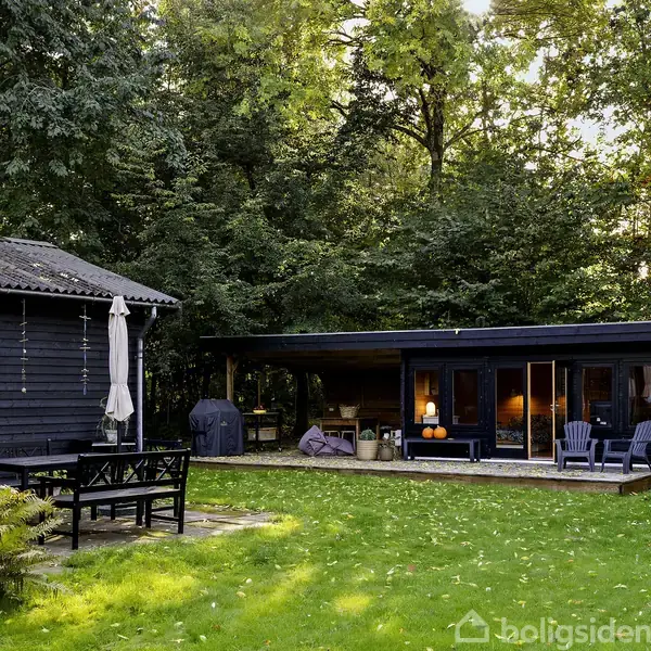 En sortmalet bygning står stille ved en terrasse omgivet af frodigt grønt græs og skov. To stole og en grill er placeret på terrassen foran bygningens glasdøre.