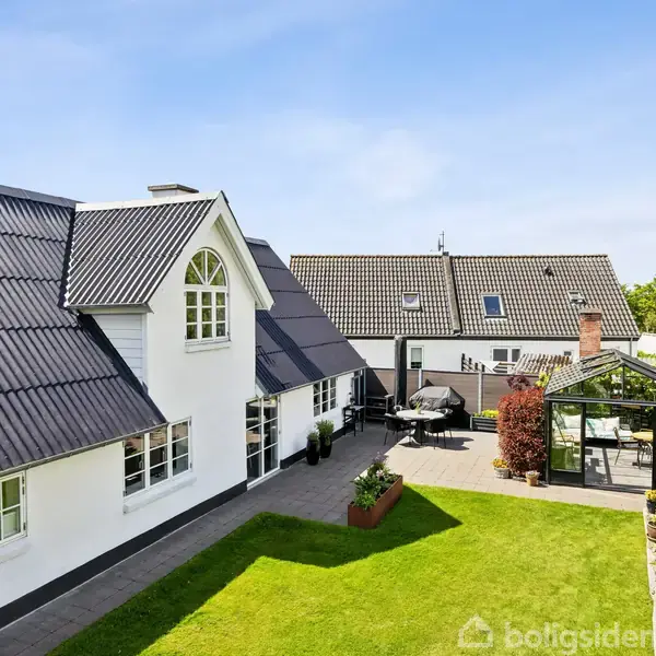 Hvidt hus med sort tag står i sollys. I haven ses en græsplæne, drivhus og terrasse med parasoller. Flere bygninger findes i baggrunden under en klar, blå himmel.