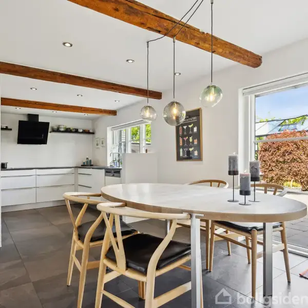 Et rundt spisebord med fire stole står i et lyst, moderne køkken med hvide skabe. Store vinduer åbner mod en terrasse og en grøn have. Der hænger tre lamper over bordet.