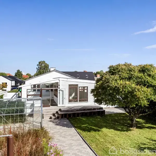 Et hvidt hus med store vinduer står i solen, omgivet af græsplæne og træer. En glasdrivhus ses ved siden af en belagt sti, der fører til huset.