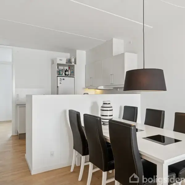 Spisebord med seks sorte stole står i et lyst køkken-alrum. En sort lampe hænger over bordet, og en hvid vase med sorte striber er placeret centralt. Bagved ses køkkenskabe.