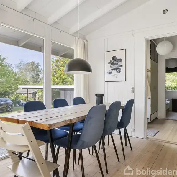 Spisebord med blå stole står i et lyst rum med store vinduer. En sort lampe hænger over bordet. Udendørs ses grønne træer og en bil i baggrunden.