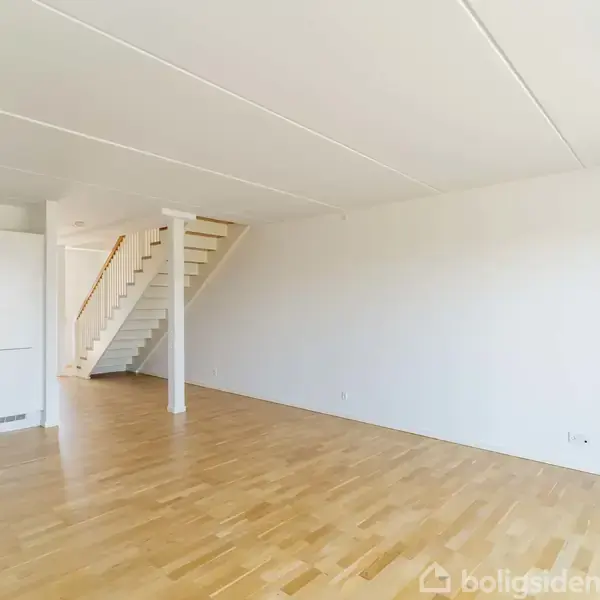 En tom stue med trægulv og hvide vægge, hvor en trappe fører opad. Der er et indbygget skab i hjørnet til venstre.