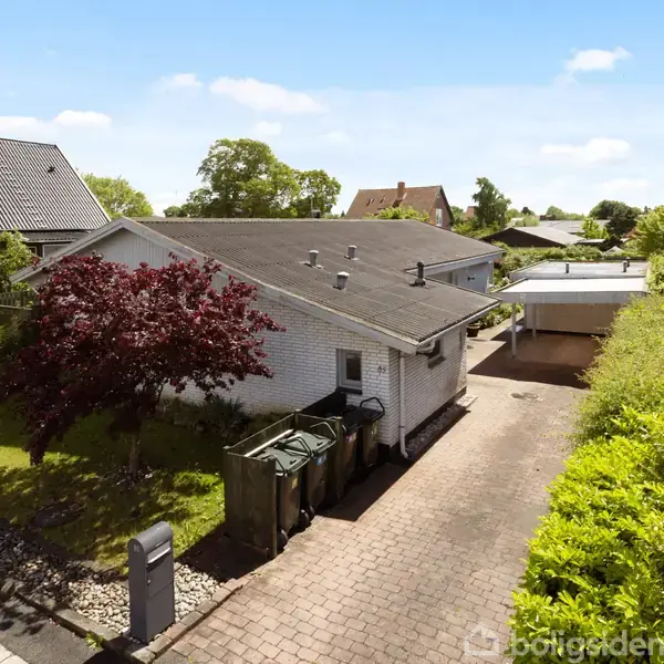 Et hvidt murstenshus med sort tag ses fra siden. Indkørslen er flisebelagt, omgivet af grønne hække og en mørkerød løn. Solrigt boligområde med flere huse i baggrunden.