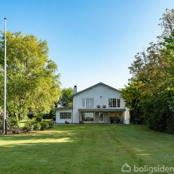 Huset står forrest midt i en velholdt have med grøn græsplæne. Det er omgivet af forskellige træer og buske. Flagstangen står til venstre. Himmel er klar og blå.