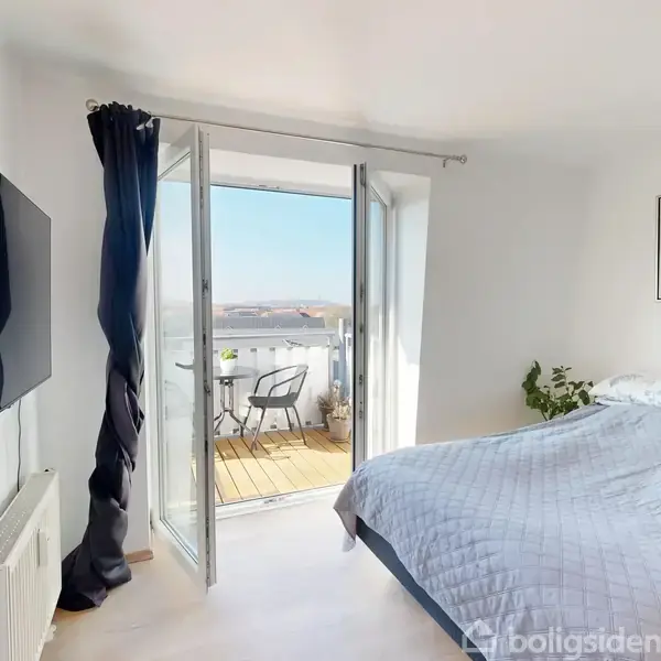 En seng står i et lyst soveværelse med åbne franske døre, der fører ud til en balkon. Der hænger et fladskærms-tv på væggen og en abstrakt kunst på den anden væg. En plante står ved siden af sengen.