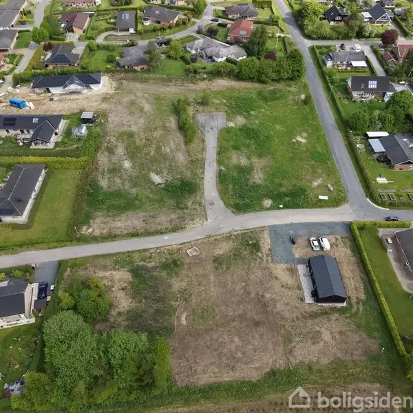 Et større, grønt område med græs og ubebyggede grunde omgivet af flere boligområder med huse. Der ses nogle indkørsler og parkerede biler i nærheden af husene.