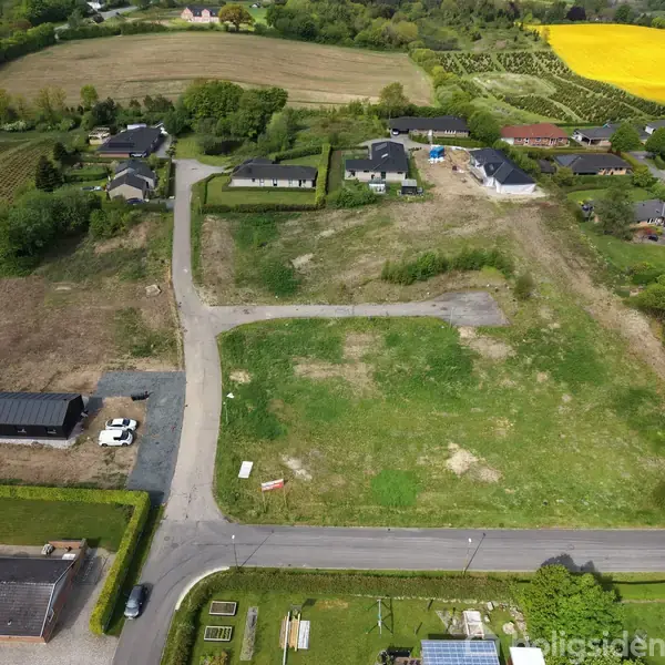 Græsklædt grund omgivet af bebyggelse; nogle biler holder parkeret ved et hus. Omgivelserne består af marker og tætliggende huse. Skilte ses på grunden.
