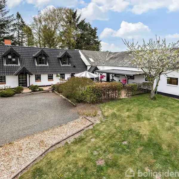 En hvidmalet landejendom med sort tag ligger i en grøn have omkranset af buske og græsplæne. Et træ blomstrer ved siden af huset. Himlen er let skyet.