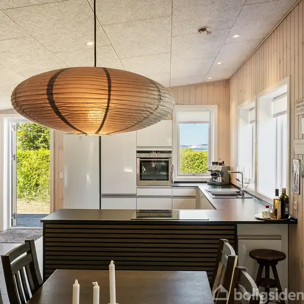 Lys træspisebord med stole under stor rund lampe. Moderne køkken i baggrunden har hvide skabe, indbygningsovn og udsigt til grøn hæk gennem vinduerne. Plakater hænger på væggen.