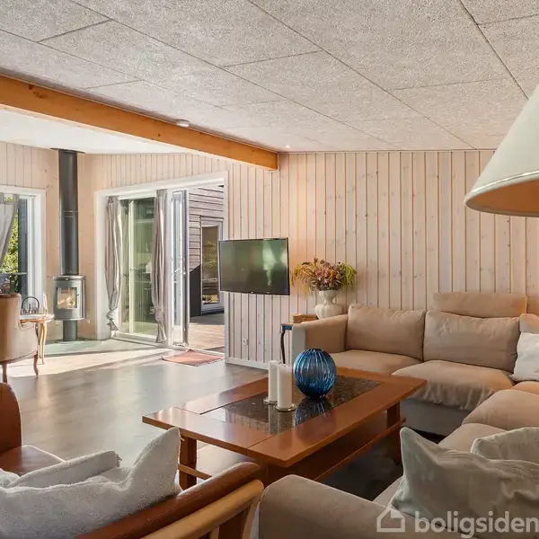 En stue med beige sofaer arrangeret omkring et sofabord, en stor lampe til højre. Pejs i baggrunden ved glasdøre fører ud til haven. Blomster på et bord og moderne indretning.