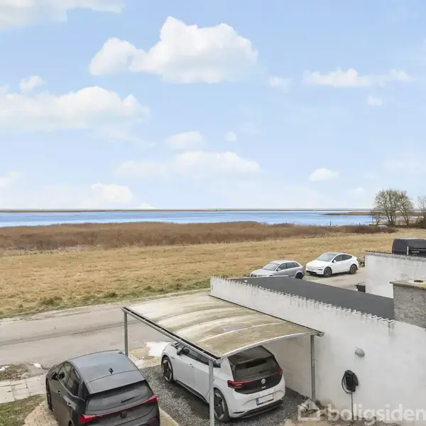 Biler holder parkeret ved en carport foran en hvid bygning. Bagved ses en vej, en græsmark og en sø i baggrunden under en blå himmel med skyer. Ingen tekst.