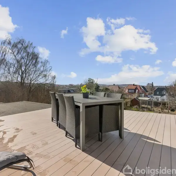 Have: Udendørs spisebord med seks stole. Aktion: Står stille på terrassen. Kontekst: Omgivet af en have med træer og udsigt til tage i nabolaget under en blå himmel med skyer.