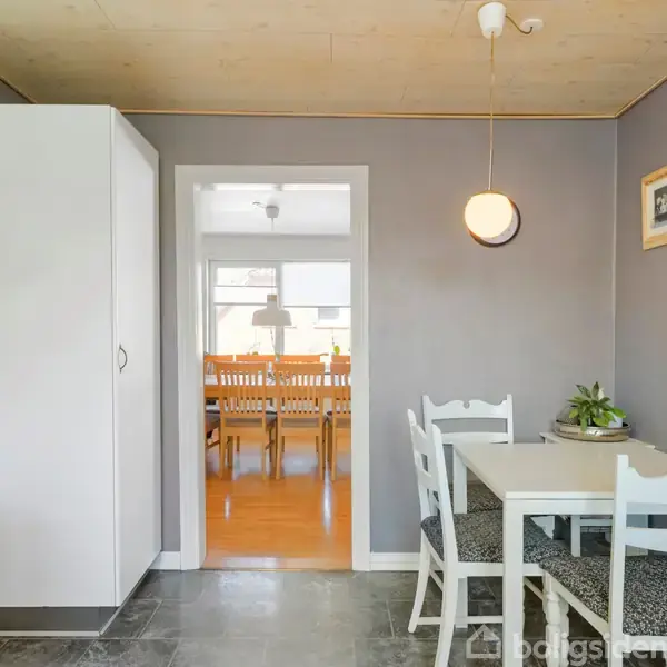 Et lille køkkenbord med to stole står i et rum med grå vægge og loftlampe. Et komfur står til venstre. Gennem en dør ses et spiseområde med træmøbler.