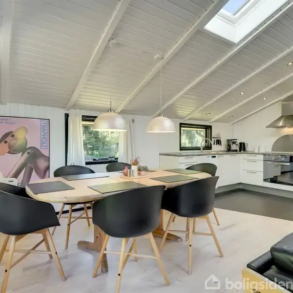 Et lyst køkken-alrum med skrå loft og ovenlysvindue. Et spisebord med seks sorte stole står centralt, omgivet af et hvidt køkken med moderne apparater. En farverig plakat hænger på væggen.
