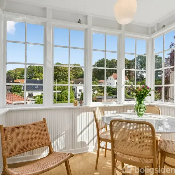 Rundt bord med blomster i vase står omgivet af baststole i en lys karnap. Store vinduer giver udsyn til træer og huse, skaber en luftig og lys atmosfære.