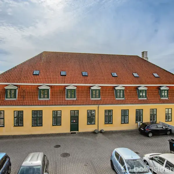 En gul murstensbygning med et rødt tegltag og mørkegrønne vinduer står stille på en brolagt gårdsplads. Flere parkerede biler omgiver bygningen under en blå, let skyet himmel.