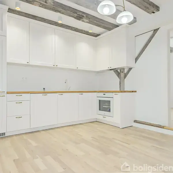 Et moderne køkken med hvide skabe og træbordplade står i et lyst rum. Synlige træbjælker præger loftet, og en stor åbning fører til en tilstødende stue.