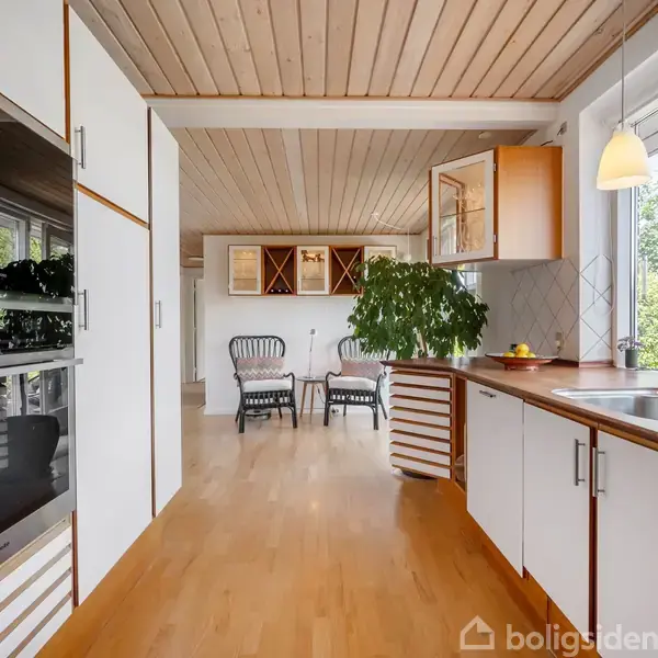 Køkken med hvide skabe og indbygget ovn, rummeligt bordplads og vask. En plante står på bordet, og tre stole er placeret ved spiseområdet. Store vinduer giver udsigt til en grøn have.