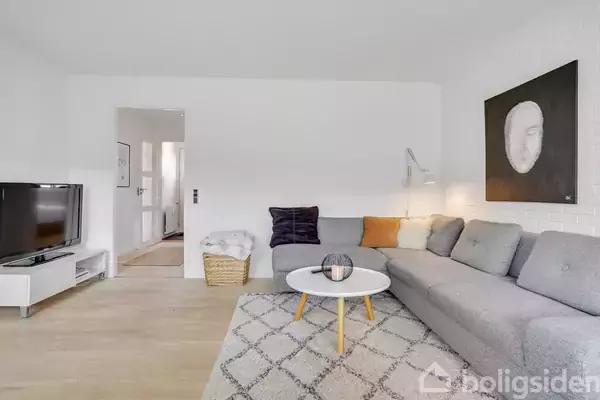 En stue med en stor grå sofa, rundt sofabord, og fladskærms-tv på et stativ. Væggene er hvide, og der hænger et maleri over sofaen. Et tæppe ligger på gulvet.