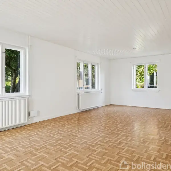 Stort, lyst rum med parketgulv, tre vinduer og to radiatorer. Udsigt til grønne træer udenfor. Væggene og loftet er hvide, og rummet virker rummeligt og luftigt.