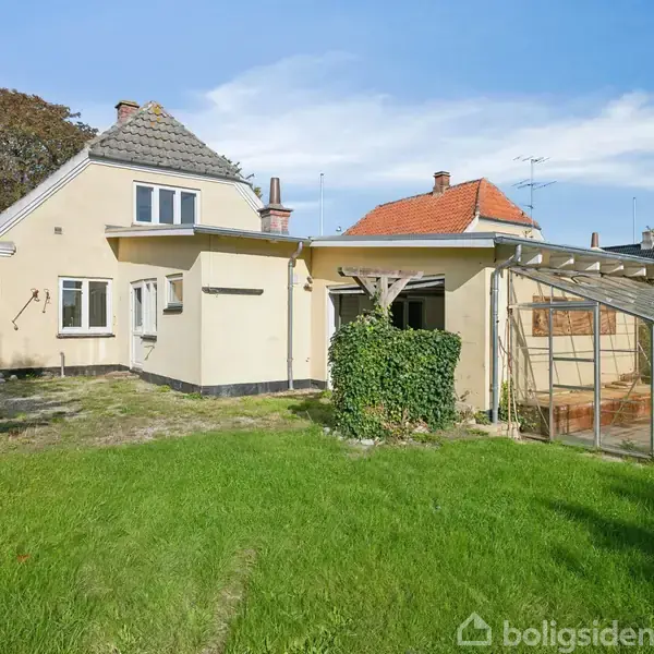 Et gult hus med en glasveranda står i en have med græsplæne og enkelte træer. Solen skinner, og himlen er blå med få skyer.