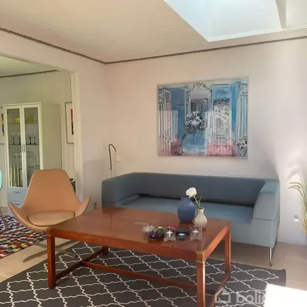 Sofa står i stue med lyse vægge. Et stort billede hænger over sofaen. Foran står et træbord med vaser. Ved siden er en lænestol og tæppe; spisestue anes i baggrunden.