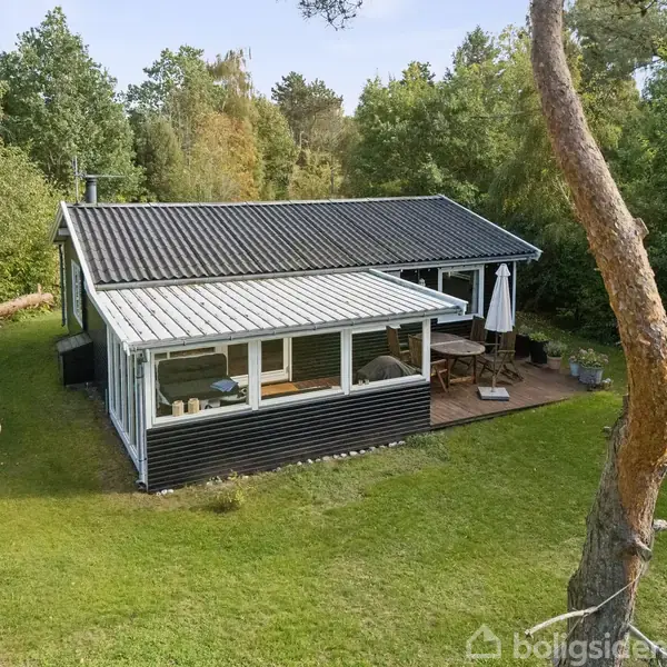 Et sort-hvidt sommerhus står omgivet af grønne træer med en overdækket terrasse udstyret med havemøbler. Huset er placeret på en velplejet græsplæne.