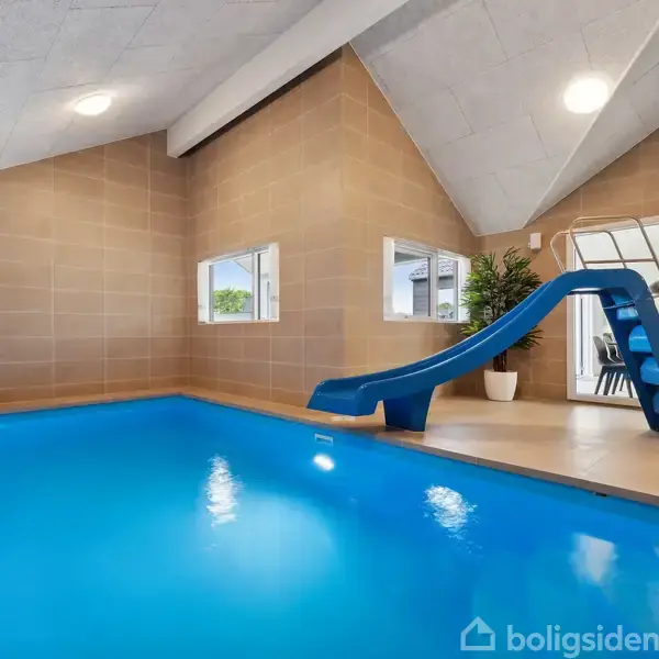 En blå vandrutsjebane fører ned i en indendørs swimmingpool. Rummet har lysebrune vægge og skråloft samt et stort vindue og grøn plante i hjørnet.