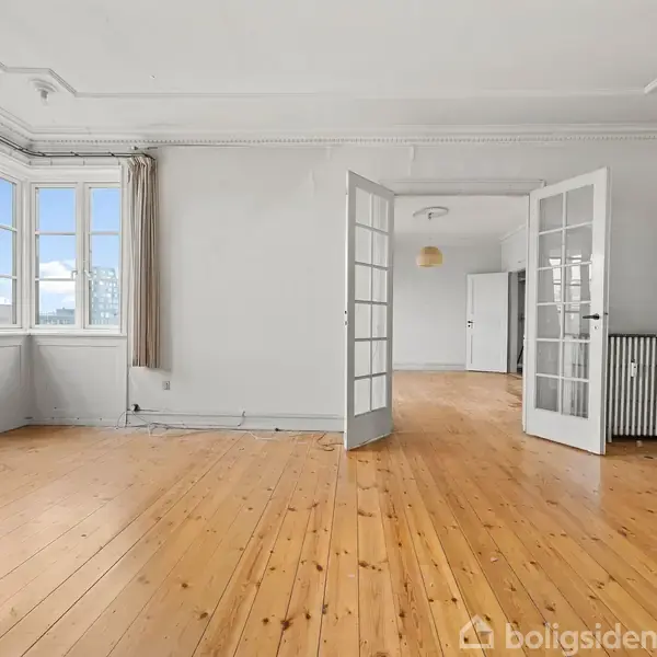 Et lyst rum med trægulv og hvide vægge, store vinduer til venstre lader lys strømme ind. To franske døre åbner ind til et tilstødende rum.