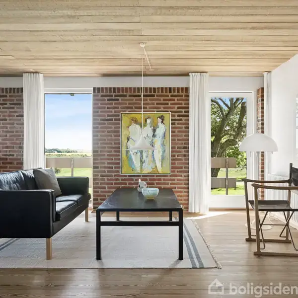 Sofaen står i en stue med træloft, murstensvægge og store vinduer. Et maleri hænger på væggen. Rummet er lyst og minimalistisk indrettet med moderne møbler.