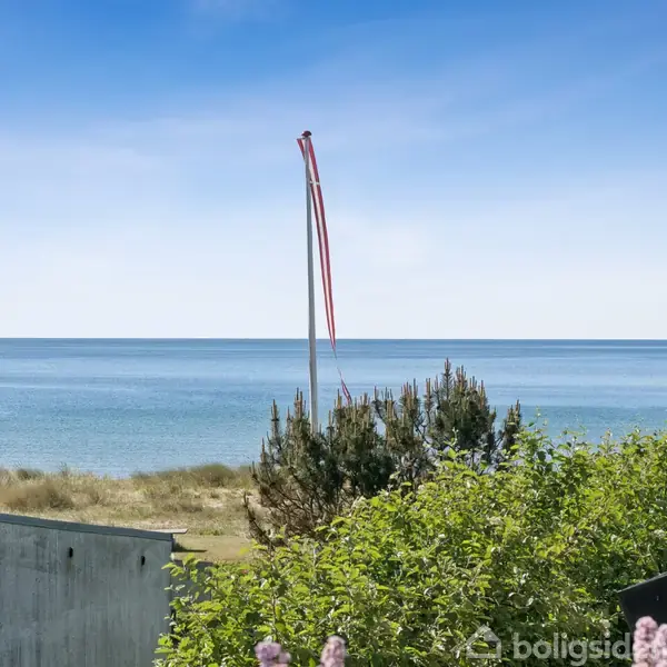 Flagstang med røde bånd står stille ved kysten. Havet ligger stilfærdigt i baggrunden, omgivet af frodige buske og træer i forgrunden.