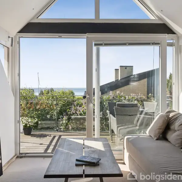 Stue med sofa og sofabord, vender ud mod terrasse med havudsigt. Terrassen har planter og udendørs møbler i solskin. Glasdøre adskiller det indendørs og udendørs område.