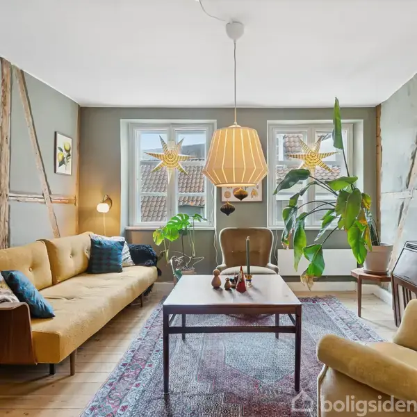 Stue med gul sofa og lænestol omkring et sofabord; en stor grøn plante står ved vinduer med stjerneudskæringer. Væggene har trædetaljer, og der hænger en lampe fra loftet.