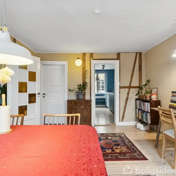 Et spisebord med rød dug står i en stue, omgivet af lysarmaturer og planter. Et skrivebord med bøger og computer er til højre. Døren fører til et andet rum.