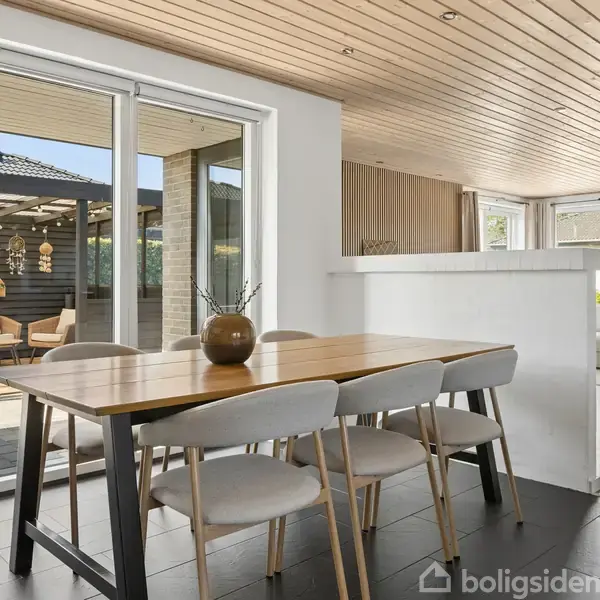 Et træbord med stole omkring står ved store glasdøre, der åbner ud til en terrasse. Boligen har et lyst, moderne interiør med trælofter og en åben stue i baggrunden.
