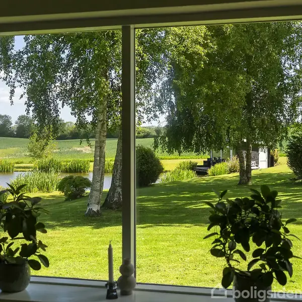 Et stort vindue viser en grøn have med træer og en lille sø i baggrunden. Indendørs står planter og stearinlys på vindueskarmen, med udsigt til en solrig dag.