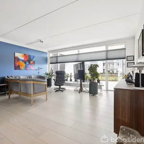 Et moderne stueområde med lyse trægulve. Der er et spisebord med stole til venstre, en farverig kunstplakat på en blå væg, og store vinduer til højre med udsigt til en altan.