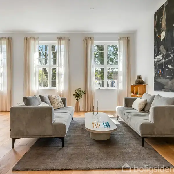 To grå sofaer står overfor hinanden omkring et ovalt sofabord med stearinlys i en lys, moderne stue med trægulv, store vinduer og et abstrakt maleri på væggen.