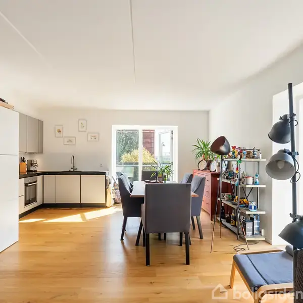 Et lyst, moderne køkkenalrum med åbent køkken og spisebord. Stort vindue fylder rummet med lys, mens en reol med planter og dekorationer står til højre. Trægulv.