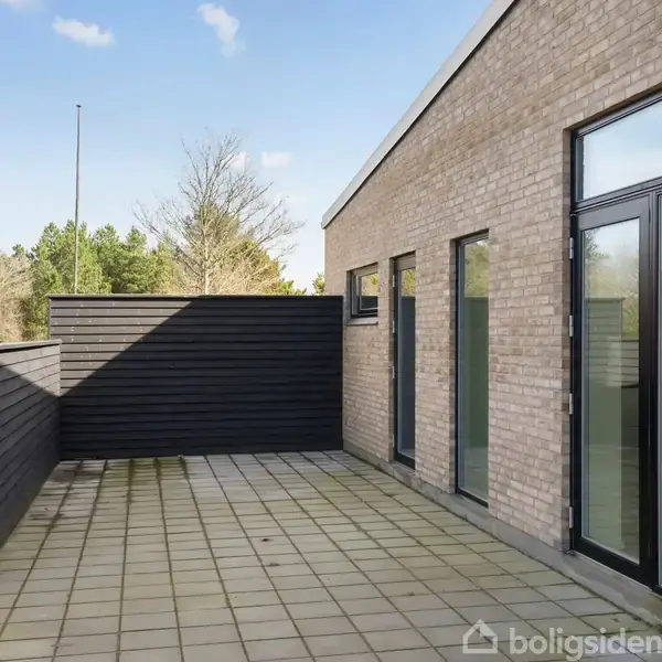 Hus af mursten med store vinduer åbner ud til en flisebelagt terrasse. Mørkt træhegn adskiller terrasse fra omkringliggende natur med træer i baggrunden. Lukket omgivelser under blå himmel.