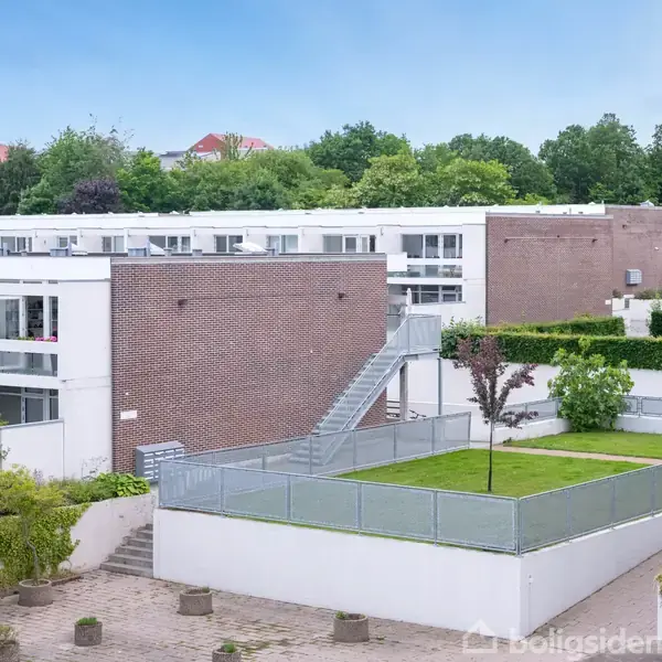 Moderne lejlighedskompleks med flade tage og store vinduer. En udendørs trappe forbinder etageadskillelserne. Omkringliggende grønt område med træer og buske giver en naturlig ramme.