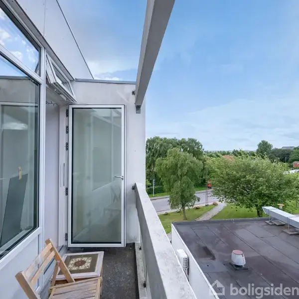 En altan med en træstol står stille ved den åbne dør. Udsigten viser en grøn park og veje under en skyet blå himmel.