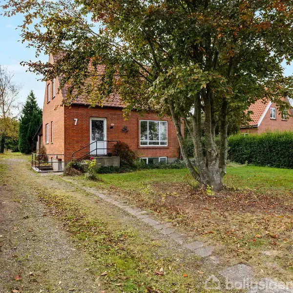 En murstensvilla med tallet "7" på facaden står langs en grussti omkranset af hække. Et stort træ vokser foran huset, der har en velholdt have.